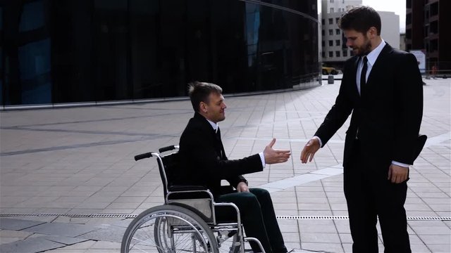 Happy boss shaking hands with disabled businessman in wheelchair outdoor in slow motion. Teal and orange style. Sunny day. Happiness. Middle shot. Modern business centre glass building background.