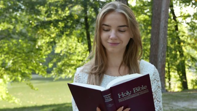 4k.Young modern girl reads  Bible in summer park. 
