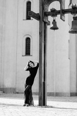 A slim, sensual brunette in a long black dress is standing by the bell tower. Fashion photo. Black-and-white photo.