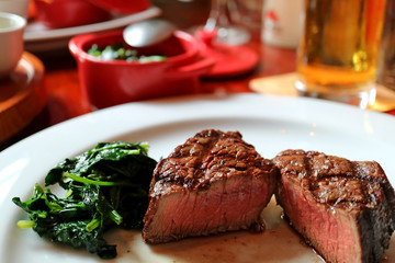 Medium grilled tenderloin steak cut in half with sauteed spinach on white plate, blurred side dishes and beverage in background  