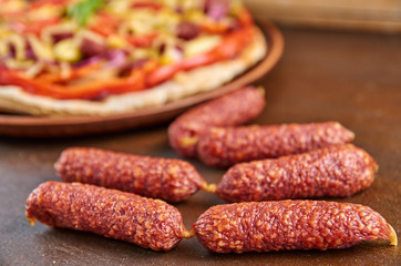Tasty little sausages salami on brown wooden table close up. On blurred background pizza with tomatoes, bell pepper, onion, green olives, corn, cheese and spices on dark plate. Side view