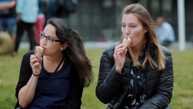 Two girls sit on the meadows in a park and eat ice cream