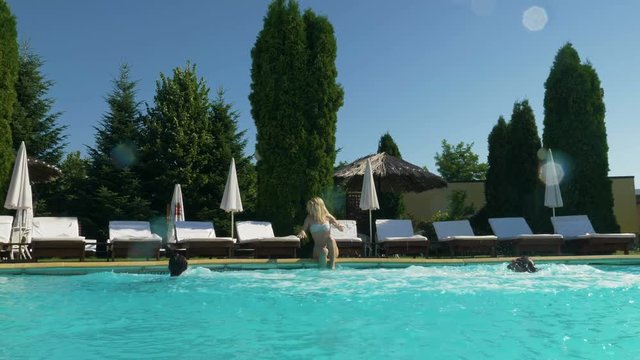 Two young men disturb young beautiful woman sitting on the edge of the pool when they jump into the pool
