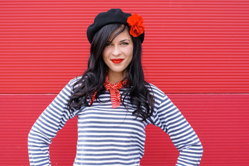 A young woman in a striped jumper, ripped jeans and beret with a flower with a red patent leather bag. A lady in French style stands opposite the red wall