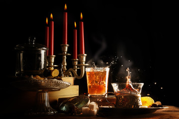 Fototapeta na wymiar hot tea with splash and steam, candles and sweets make the dark wintertime cozy, still life against a black background with copy space