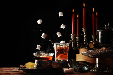 sugar cubes falling into teacups of glass on a rustic table setting with candles and candies, cozy sweet break in the dark winter time, black background with copy space, selective focus