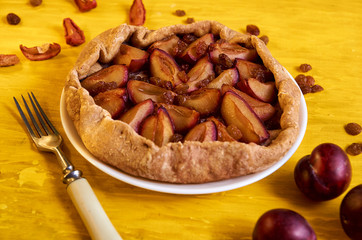 Plums pie with raisins on a white plate on yellow wooden background decorated with three fresh plums, brown raisins, dried pieces of apple and silver vintage fork. Side view