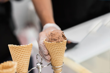 Obraz na płótnie Canvas Waffle cone, horn with ball of chocolate, coffee soft ice cream and hand of seller. Delicious cooling portion for fun. Selective focus. Real scene in store