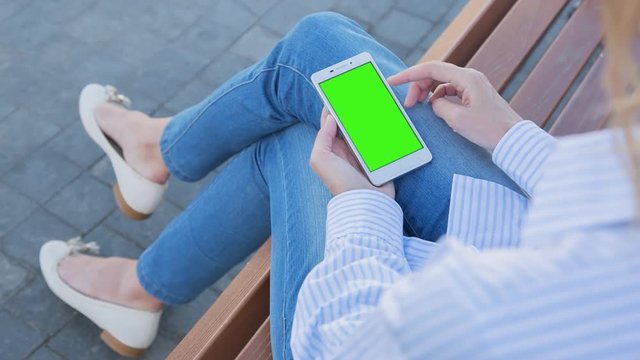 Young Woman in blue jeans sitting on bench uses SmartPhone with pre-keyed green screen. Few types of gestures - scrolling up and down, tapping, zoom in and out. Perfect for screen compositing
