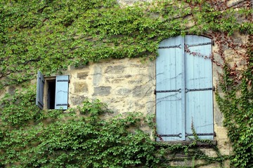 One small and one large pair of pale blue wooden shutters on an on stone French house overgrown...