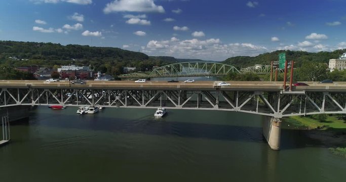 A reverse aerial wide shot view of traffic traveling on a bridges in Western Pennsylvania while a recreational boats pass underneath.  	