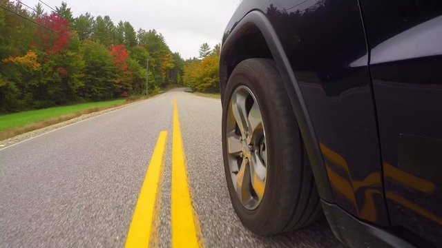 CLOSE UP Black SUV car driving along double yellow line road past colorful trees on autumn day. Detail of car tire spinning while driving through bright autumn forest in sunny fall. Car tyre rolling