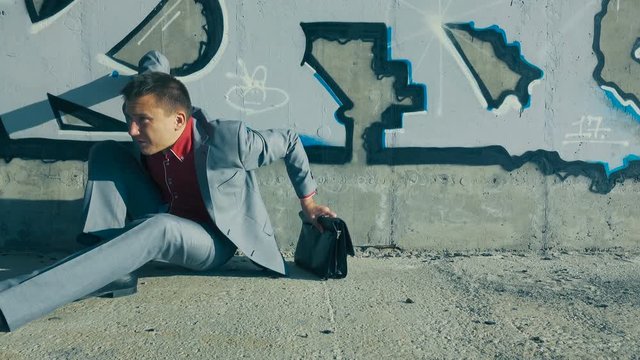 Unemployed businessman in a suit sleeps near a concrete wall, putting a briefcase under his head