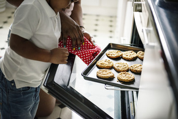 Black kid helping mom baking cookies in the kitchen - obrazy, fototapety, plakaty