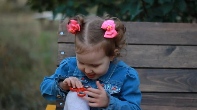 Funny cute little girl is playing with a spinner in the park on the bench, shows focus and twists the spiner on the knee.