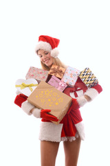 A young girl dressed in a Santa Claus suit has lots of Christmas presents gathered in her arms. Smiling, she stands in front of a white background.