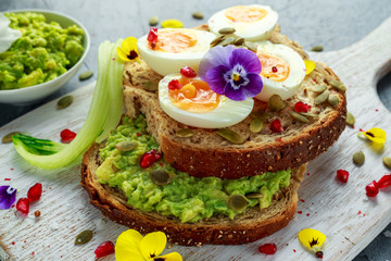 Tasty sandwich with avocado boiled eggs, pumpkin seed and edible viola flowers in a white board. healthy food