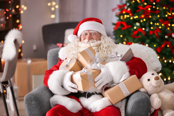 Fototapeta na wymiar Authentic Santa Claus with gift boxes sitting in armchair at room decorated for Christmas