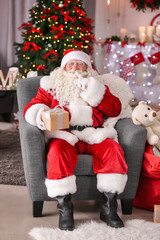 Authentic Santa Claus with gift box sitting in armchair at room decorated for Christmas