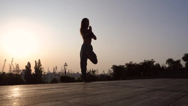 Young beautiful caucasian woman doing yoga asana on stairs in the city. Summer morning. Girl wearing blue pants, black top and mala beads for meditation. Surya Namaskar. Slow motion.