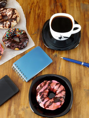 Doughnut with pink glazing, cup of coffee and stationery on a wooden table. Morning of a business woman