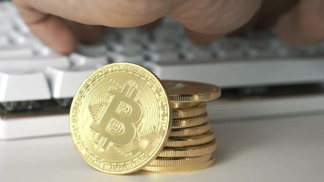 A stack of bitcoin coins while a hand is typing on a computer keyboard in the background. Bitcoin is a worldwide cryptocurrency and digital payment system.