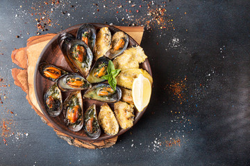 Overhead of seafood dinner table. Grilled baked mussels with butter, parsley and cheese on ceramic plate on dark background. Top view.