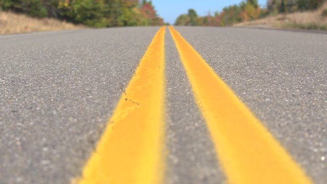 CLOSE UP DOF: Double yellow lines on empty interstate road trough autumn forest on sunny day. Brand new twin yellow lines on empty highway road past colorful trees. Yellow doubles on road surface