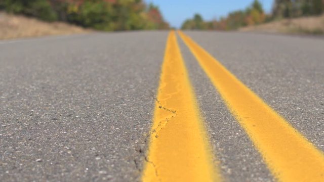 CLOSE UP DOF: Double yellow lines on empty interstate road trough autumn forest on sunny day. Brand new twin yellow lines on empty highway road past colorful trees. Yellow doubles on road surface