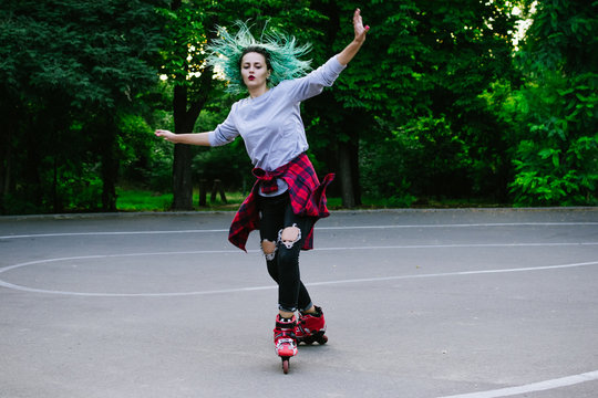Young stylish funky girl with green hair riding roller skates and dancing in park