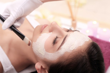 Obraz na płótnie Canvas Beautiful young woman is getting facial clay mask at spa, lying with cucumbers on eyes
