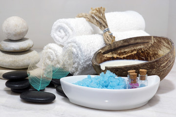 A set for Spa treatments with coconut milk, hot stones and blue bath salt is located on a white marble countertop.