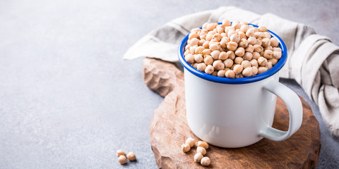 Raw uncooked organic chickpeas in white enamel mug on rustic wooden board on light gray stone background. Healthy vegan vegetarian food concept. Banner.