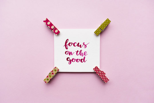 Inspirational quote "Focus on the good" handwritten with watercolor in calligraphy style, miniature clothespins on a pink background. Flat lay
