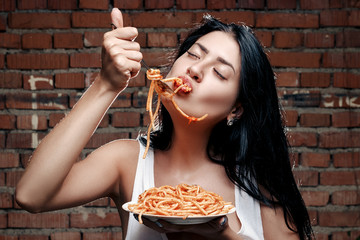 Sexy seductive cheeky girl in a white T-shirt and a plate of pasta and a fork wildly eats pasta with ketchup standing in glasses on the background of a brick wall - Powered by Adobe
