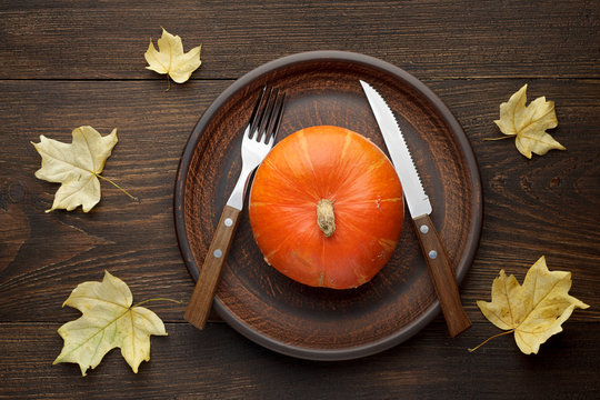 Autumn table setting with pumpkin and cutlery on plate on wooden background with maple leaves.