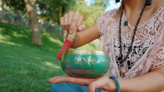 Woman playing singing bowl while sitting on pink yoga mat in park at summer. Vintage tonned. Beautiful girl with mala beads meditating on green grass. Slow motion.