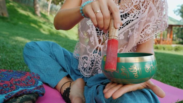 Woman playing singing bowl while sitting on pink yoga mat in park at summer. Vintage tonned. Beautiful girl with mala beads meditating on green grass. Slow motion.