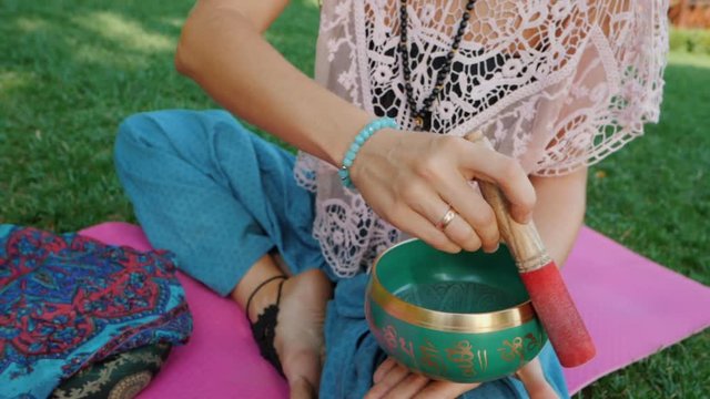 Woman playing singing bowl while sitting on pink yoga mat in park at summer. Vintage tonned. Beautiful girl with mala beads meditating on green grass. Slow motion.