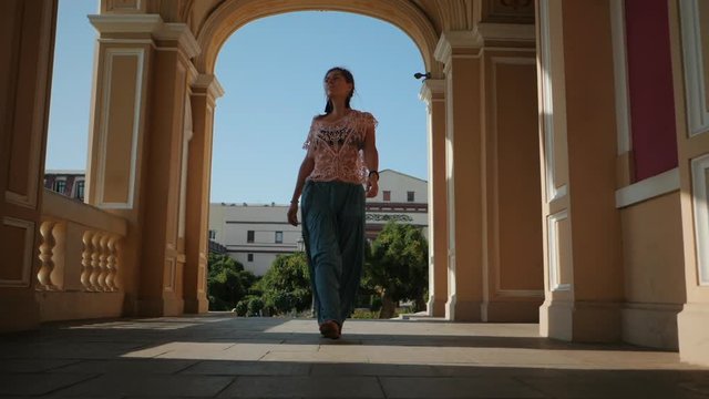Beautiful girl in blue pants and pink lace top walking in historic architecture. Yoga in the city. Health concept. Slow motion. Woman goes to the camera and smiling.