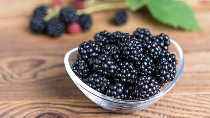 Sweet juicy blackberries in a glass bowl. Fresh forest fruits on wooden table with blurred bramble branch in the background. HD ratio 16x9.