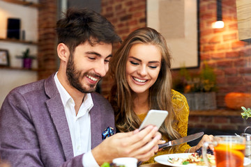 Happy cute young couple looking at phone and smiling while eating and sitting in a cafeteria
