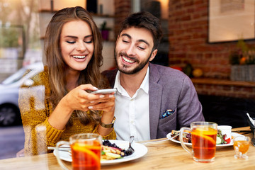 Young loving couple looking at phone and taking photos of their breakfast while sitting together in a cafeteria