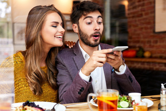 Cute cheerful young couple having fun while sitting in a restaurant, taking photos while eating and smiling
