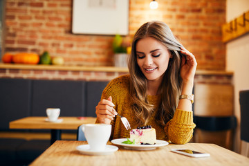 Portrait of a gorgeous young lady smiling while eating cake sitting in a cafeteria