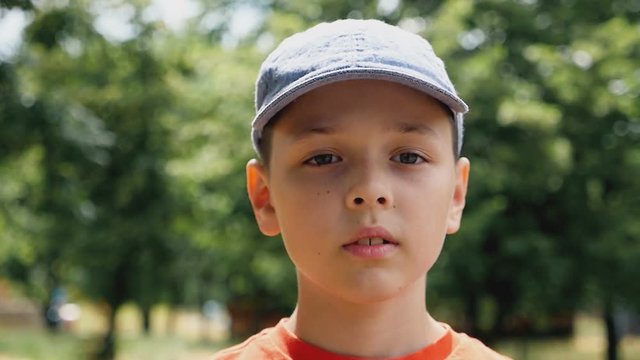 A thoughtful seven-year-old boy with deep eyes and a cap on his head stands in a park on a picturesque lawn and thinks about his life with on a sunny day in summer in slow motion