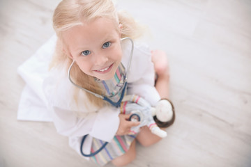 Little girl in medical coat playing with doll on floor