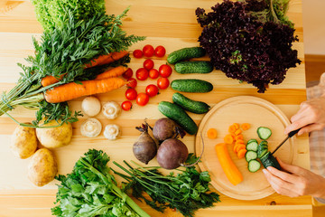 Vegetable on the wooden table in the kitchen - woman's hands cutting cucumber on wooden board in kitchen. Top view.
