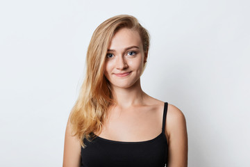 Fototapeta premium Adorable blue eyed female with long straight light hair, wears casual black t-shirt, looks happily into camera as going to walk with boyfriend, has good mood, isolated over white studio background