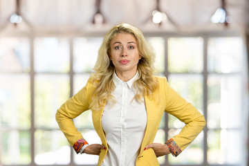 Serious young blonde holding hands on hips. Confident young business woman posing with hands on hips on blurred background.